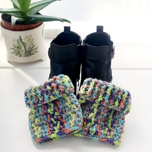 Crochet boot cuff ankle warmers made by Sarah Lou Crafts, in a multicoloured yarn called Fireworks. Shown laying on black ankle boots with the top cuff folded over to been as a short option.