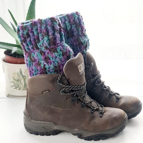 Crochet boot cuffs made by Sarah Lou Crafts, in a purple colourway. Shown in brown Scarpa Hiking Boots on a white table with green aloe vera plant in the back ground.