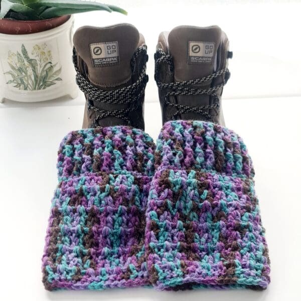 Crochet ankle warmer boot cuffs made by Sarah Lou Crafts, in a purple colourway Shown laying on brown hiking boots on a white table with green aloe vera plant in the background.