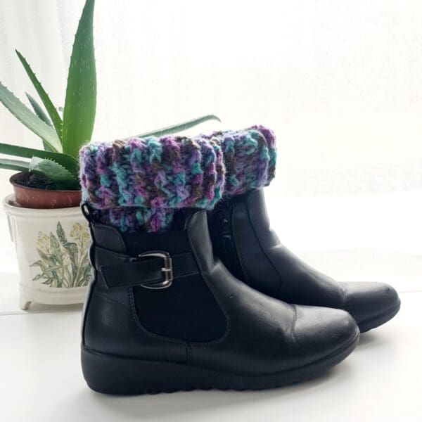 Crochet boot cuffs made by Sarah Lou Crafts, in a purple colourway Shown in black ankle boots on a white table with green aloe vera plant in the background.