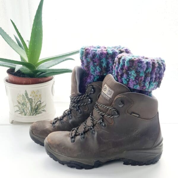 Crochet boot cuff ankle warmers made by Sarah Lou Crafts, in a purple colourway Shown in brown boots as a short option by folding the top down, on a white table with green aloe vera plant in the background.