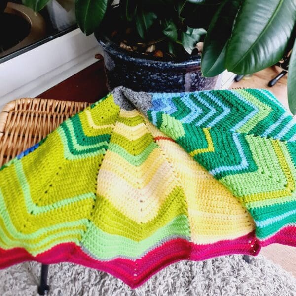 Multicoloured crochet blanket by Sarah Lou Crafts, shown folded laying on a brown wicker foot stool.