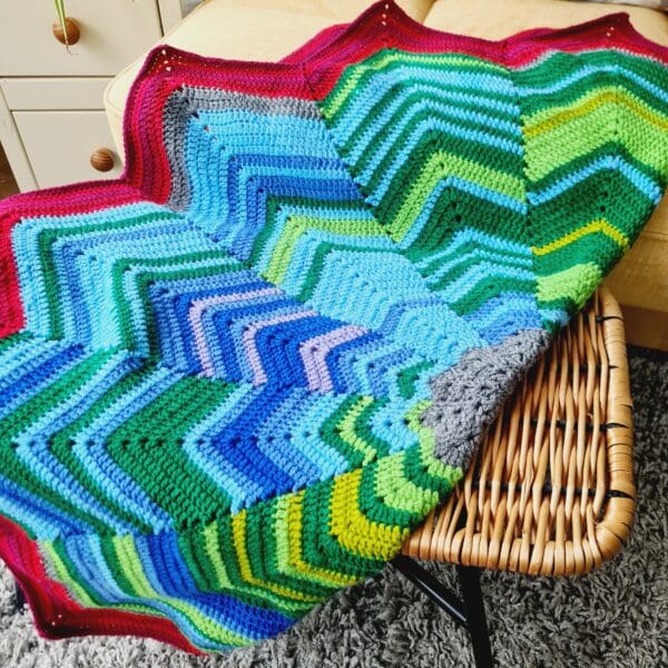 Multicoloured crochet blanket by Sarah Lou Crafts, shown folded in half across a brown wicker foot stool.