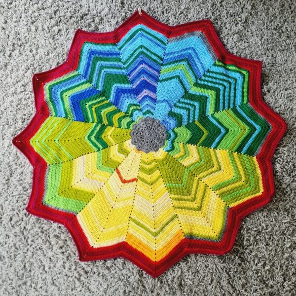 Rainbow crochet star blanket by Sarah Lou Crafts, shown spread out flat on a grey fluffy rug.