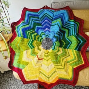 Crochet star blanket made by Sarah Lou Crafts using a rainbow of DK yarns to create a 2024 temperature blanket. Each segment of the 12 point star is one month of daily top temp taken at 12 pm. Shown draped on a yellow sofa.