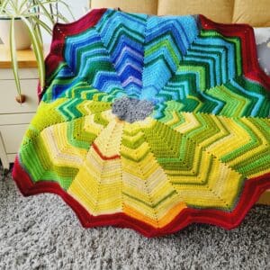 Multicoloured crochet star shaped blanket by Sarah Lou Crafts, shown draped on a yellow sofa.