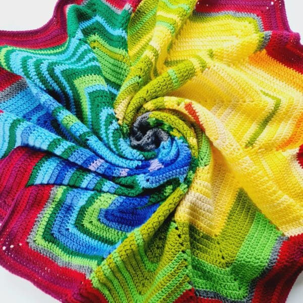 Multicoloured crochet blanket by Sarah Lou Crafts, shown in a twisted spiral on a white table. Photo taken from above looking down.