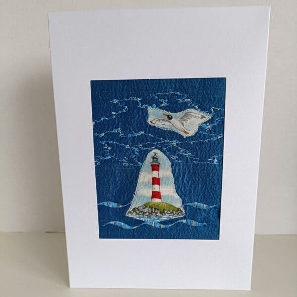 Front of card made from collage of textiles with machine stitching showing coastal view of lighthouse with sea gull on blue silk background.