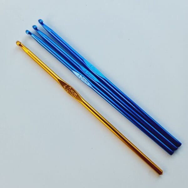 Three blue coloured metal crochet hooks and one gold coloured metal hook, all 4 mm size, laying diagonally on a white table.