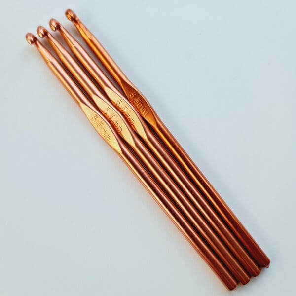 Three rose gold coloured metal crochet hooks, size 5,5 mm, laying diagonally on a white table.