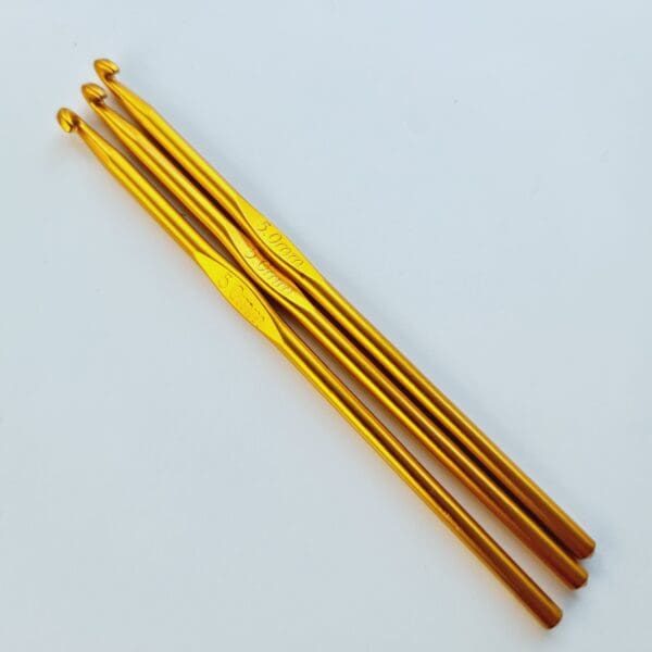 Three gold coloured metal crochet hooks, 5 mm size, laying diagonally on a white table.