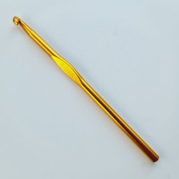 Single gold coloured metal crochet hook, 6 mm size, laying diagonally on a white table.