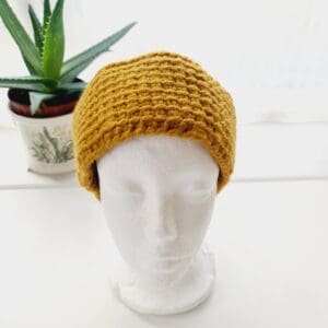 Gold coloured chunky ear warmer headband made by Sarah Lou Crafts using a Tunisian Crochet technique. Shown front facing on a white mannequin head with an aloe vera plant in the background.