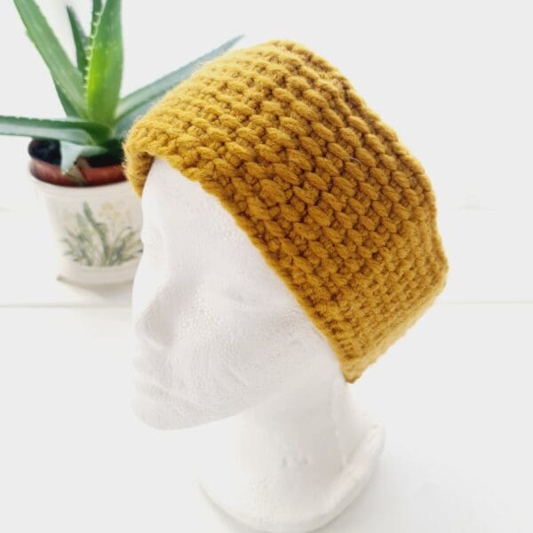 Angled view of a chunky gold ear warmer headband made by Sarah Lou Crafts, shown on a white mannequin head facing slightly to the left, with an aloe vera plant in the background.