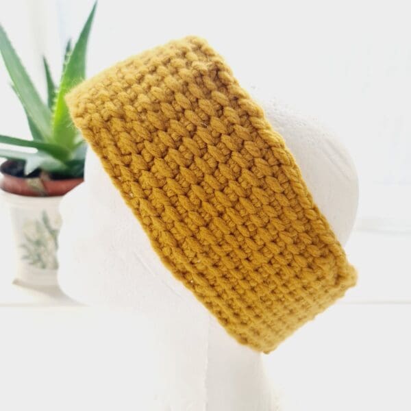 Side view of a chunky gold ear warmer headband made by Sarah Lou Crafts, shown on a white mannequin head facing to the left, with an aloe vera plant in the background.