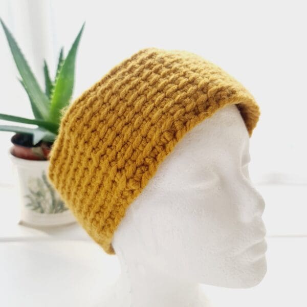 Angled view of a chunky gold ear warmer headband made by Sarah Lou Crafts, shown on a white mannequin head facing slightly to the right, with an aloe vera plant in the background.