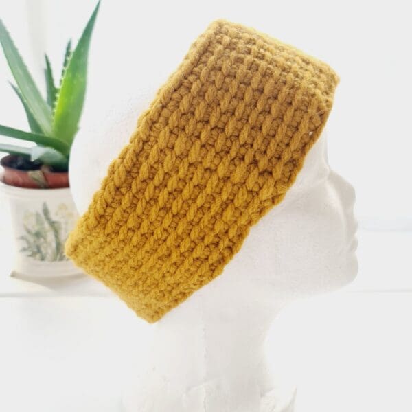 Side view of a chunky gold ear warmer headband made by Sarah Lou Crafts, shown on a white mannequin head facing to the right, with an aloe vera plant in the background.