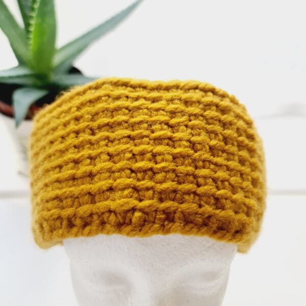 Chunky gold ear warmer headband made by Sarah Lou Crafts using a Tunisian Crochet technique. Shown on a white mannequin head with an aloe vera plant in the background.