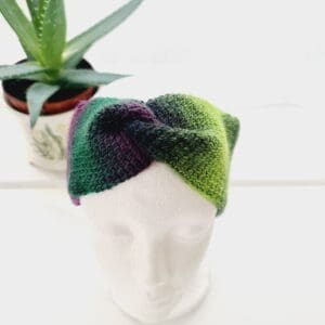 Purple and green coloured crochet ear warmer headband made by Sarah Lou Crafts using a Tunisian Crochet technique. Shown front facing on a white mannequin head with an aloe vera plant in the background.