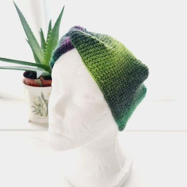 Angled view of a green and purple ear warmer headband made by Sarah Lou Crafts, shown on a white mannequin head facing slightly to the left, with an aloe vera plant in the background.