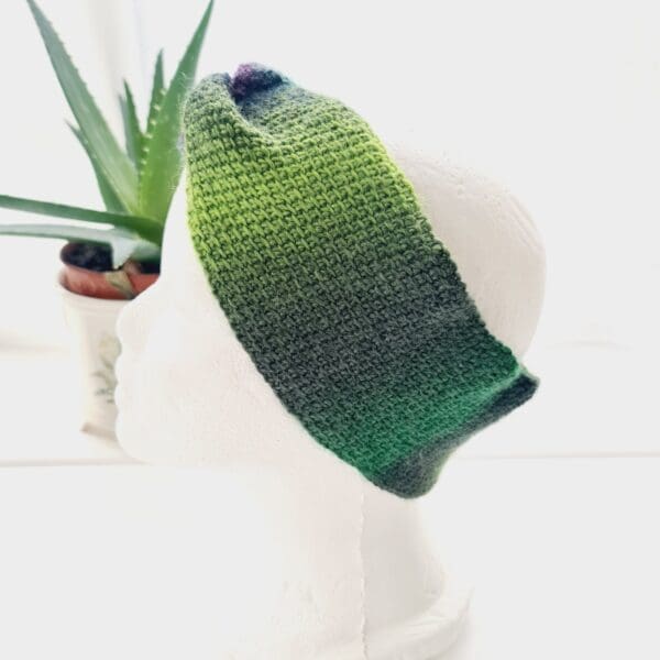 Side view of a green and purple crochet ear warmer headband made by Sarah Lou Crafts, shown on a white mannequin head facing to the left, with an aloe vera plant in the background.