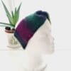 Angled view of a purple and green ear warmer headband made by Sarah Lou Crafts, shown on a white mannequin head facing slightly to the right, with an aloe vera plant in the background.