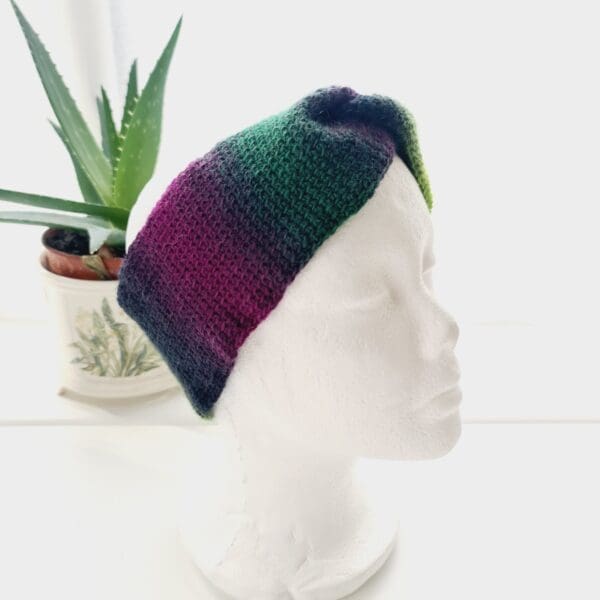 Angled view of a purple and green ear warmer headband made by Sarah Lou Crafts, shown on a white mannequin head facing slightly to the right, with an aloe vera plant in the background.