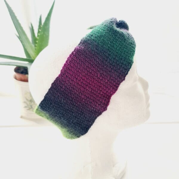 Side view of a purple and green ear warmer headband made by Sarah Lou Crafts, shown on a white mannequin head facing to the right, with an aloe vera plant in the background.