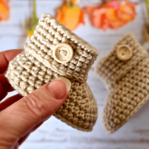 a pair of parchment coloured crochet baby booties, with a neat folded cuff and finished with a handmade with love button