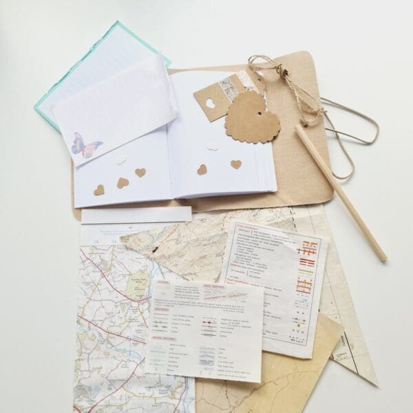 Flat lay photo showing components of a Junk Journal Starter Kit by Sarah Lou Crafts. Kit includes, Rainbow Notebook, Pencil, Map Scraps, Gift Tags, Washi Tape & String.