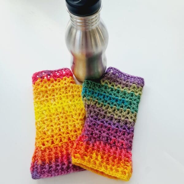 Rainbow crochet mitts, made by Sarah Lou Crafts. Shown laying flat on a white table with a silver metal water bottle nearby.