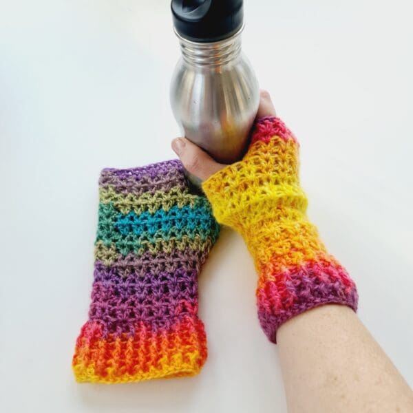 Rainbow crochet mitts, made by Sarah Lou Crafts. Shown being worn while holding a silver metal water bottle.