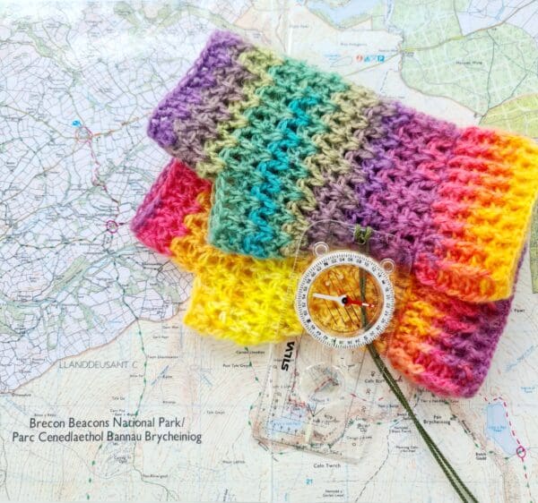 Rainbow crochet fingerless gloves, made by Sarah Lou Crafts, shown laying on top of a Brecon Beacons maps with a compass to the side.