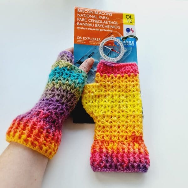 Rainbow fingerless mitts, made by Sarah Lou Crafts, in a soft variegated yarn. Shown being worn while holding a map and compass.