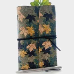 Cork fabric notebook journal with green and gold leaves pattern. Photographed standing again a green plant with silver pen in front.