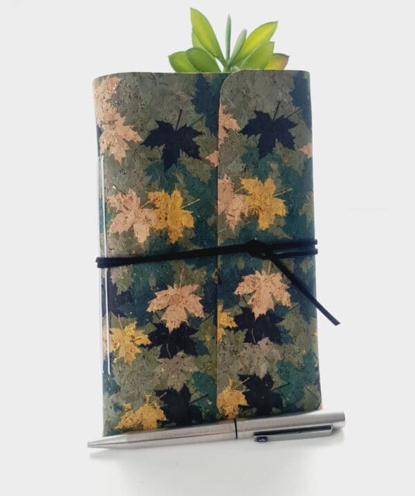 Cork fabric notebook journal with green and gold leaves pattern. Photographed standing again a green plant with silver pen in front.