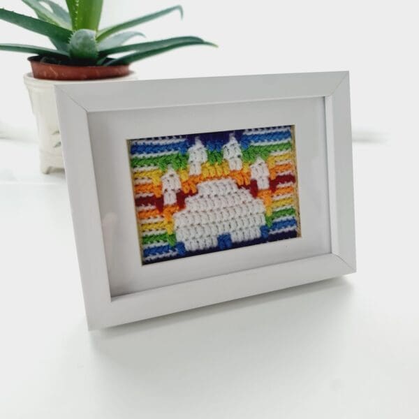 Framed crochet picture of a dog's paw print in white with a rainbow stripe background, shown in a white box frame.