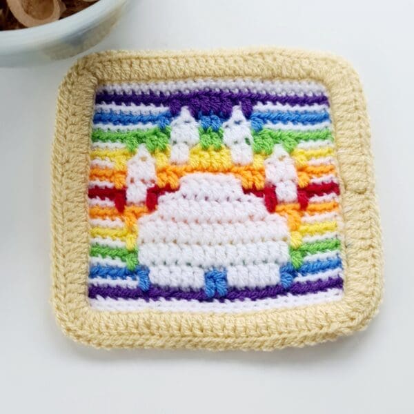 Close up photo of a white paw print mat with a rainbow background and cream border made by Sarah Lou Crafts, shown laying flat on a white table.