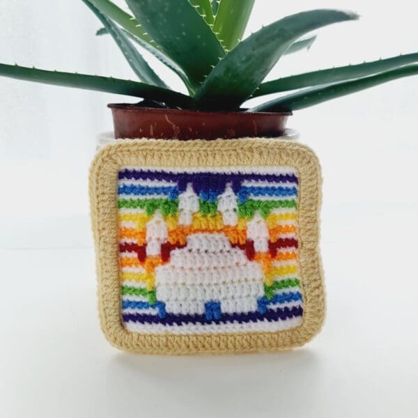 Mosaic Crochet mat with a white paw print and rainbow background made by Sarah Lou Crafts, shown stood against a small potted plant.