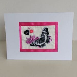 Textile collage of butterfly with 3 heart-shaped buttons and sparkly beads surrounded with pink ribbon border and set in white frame of card.