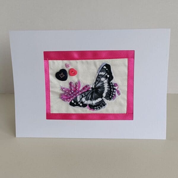 Textile collage of butterfly with 3 heart-shaped buttons and sparkly beads surrounded with pink ribbon border and set in white frame of card.