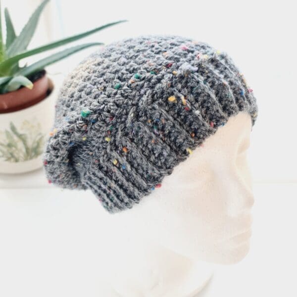 Slate grey crochet slouchy beanie hat with wide brim shown on a white mannequin head facing to the left with an aloe vera house plant in the background.