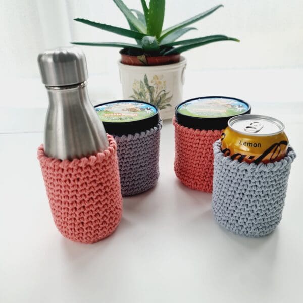 Group photo of four crochet cotton stubby holders, made by Sarah Lou Crafts, 2 pink and 2 grey for showing with ice cream tubs, metal bottle and drinks can in each.