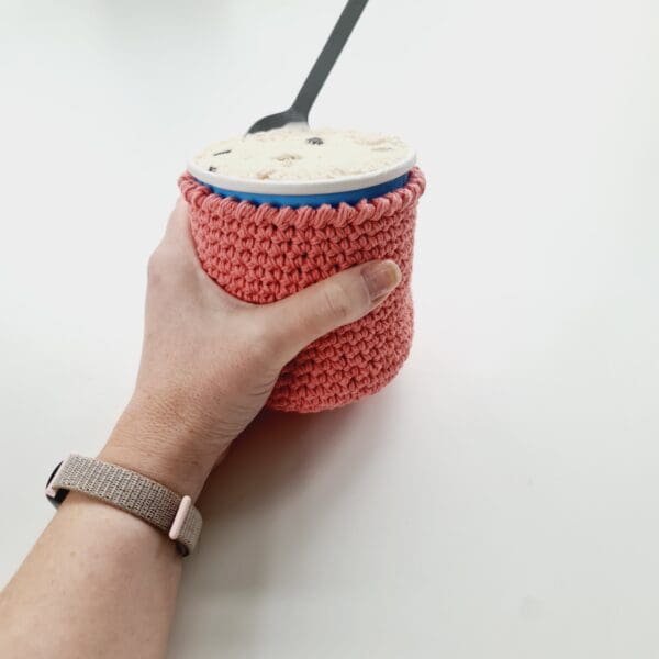 Pink crochet cotton stubby holder for round ice cream tub, made by Sarah Lou Crafts, shown being held with and open tub of ice cream and spoon sticking out.