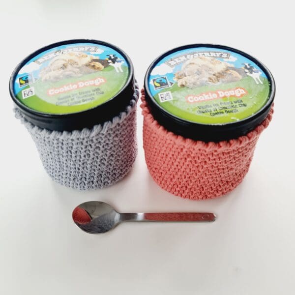 Pair of crochet cotton stubby holders for round ice cream tubs, made by Sarah Lou Crafts, one each in grey and pink holding 2 tubs of Ben & Jerry's with a small silver teaspoon in front.