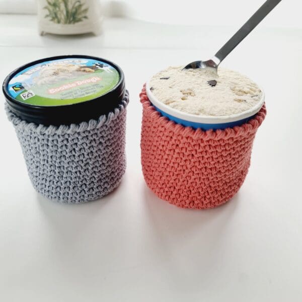 Pair of crochet cotton stubby holders for round ice cream tubs, made by Sarah Lou Crafts, one each in grey and pink holding 2 tubs of Ben & Jerry's.