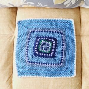 Crochet square in a moss stich pattern with blue and lavender rows showing the temperatures for January. Shown laying flat on a yellow sofa. 