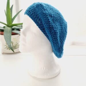 Close-up of a teal sea blue coloured crochet beret. The intricate stitch pattern creates a raised, textured surface. The yarn is soft and appears thick, ideal for spring outfits and summer festival wear.