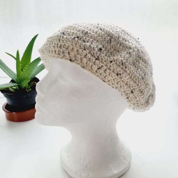 A white mannequin head wearing a cream crochet beret, by Sarah Lou Crafts. The beret is styled slightly to the side, framing the face.