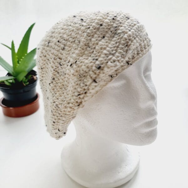 Crochet winter beret made by Sarah Lou Crafts using premium acrylic yarn in a cream colour with darker flecks throughout, called Berber. Shown on white mannequin head with plaint in back ground.